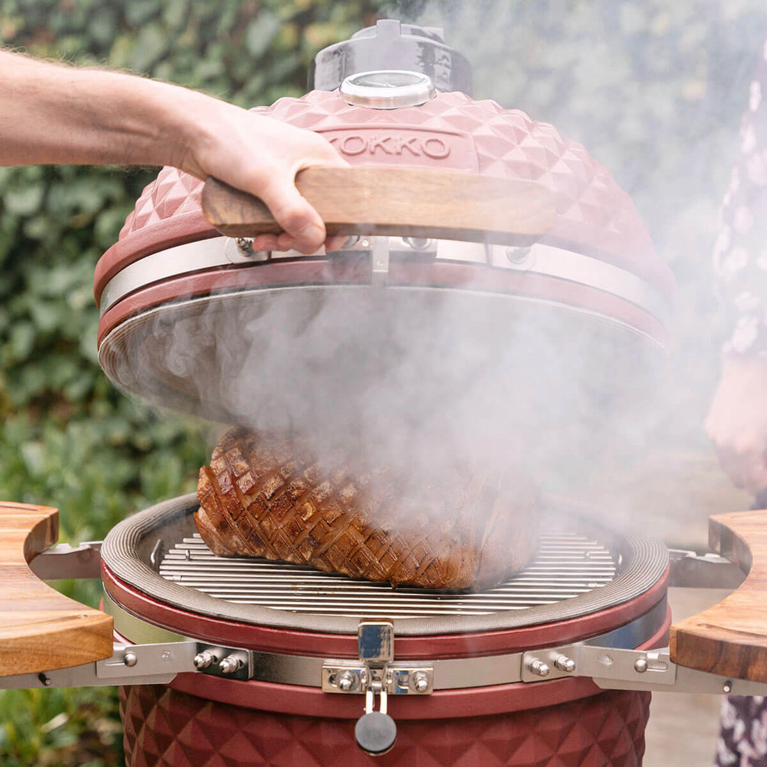 Eine Hand hebt den Deckel eines roten Kokko Kamado an und gibt den Blick auf ein gegrilltes Stück Fleisch auf dem Edelstahlrost frei. Dichte Rauchschwaden steigen aus dem Korpus auf und unterstreichen das Kocherlebnis mit Holzfeuer, die umlaufende Dichtung sowie die seitlichen Ablagen aus Holz.