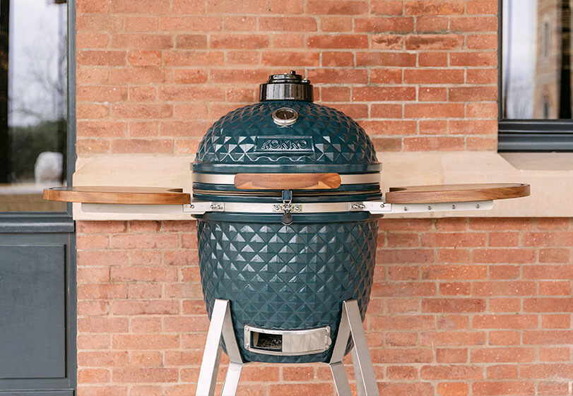 Kamado Kokko in Ozeanblau auf Gestell mit Holzablagen, frontal im Freien vor einer Ziegelmauer präsentiert.