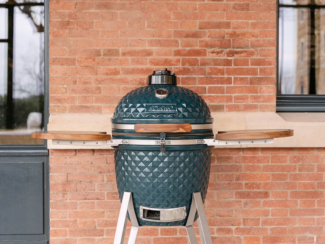 Kamado Kokko in Ozeangrün, im Außenbereich vor einer Ziegelmauer aufgestellt. Der Grill ist geschlossen, mit einem strukturierten Keramikdeckel, integriertem Thermometer auf der Oberseite, Holzgriff, Sicherheitsverschluss aus Edelstahl und zwei seitlichen Ablagen aus Akazienholz. Das Ganze steht auf einem Metallgestell mit sichtbarem unterem Lüftungsbehälter.