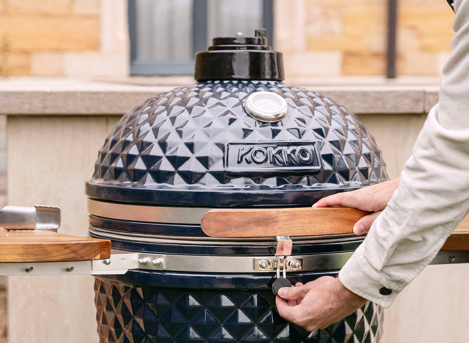 Hand betätigt den Sicherheitsriegel eines schwarzen Kokko Kamado mit texturiertem Keramikdeckel, Holzgriff und integriertem Thermometer, im Einsatz im Freien.