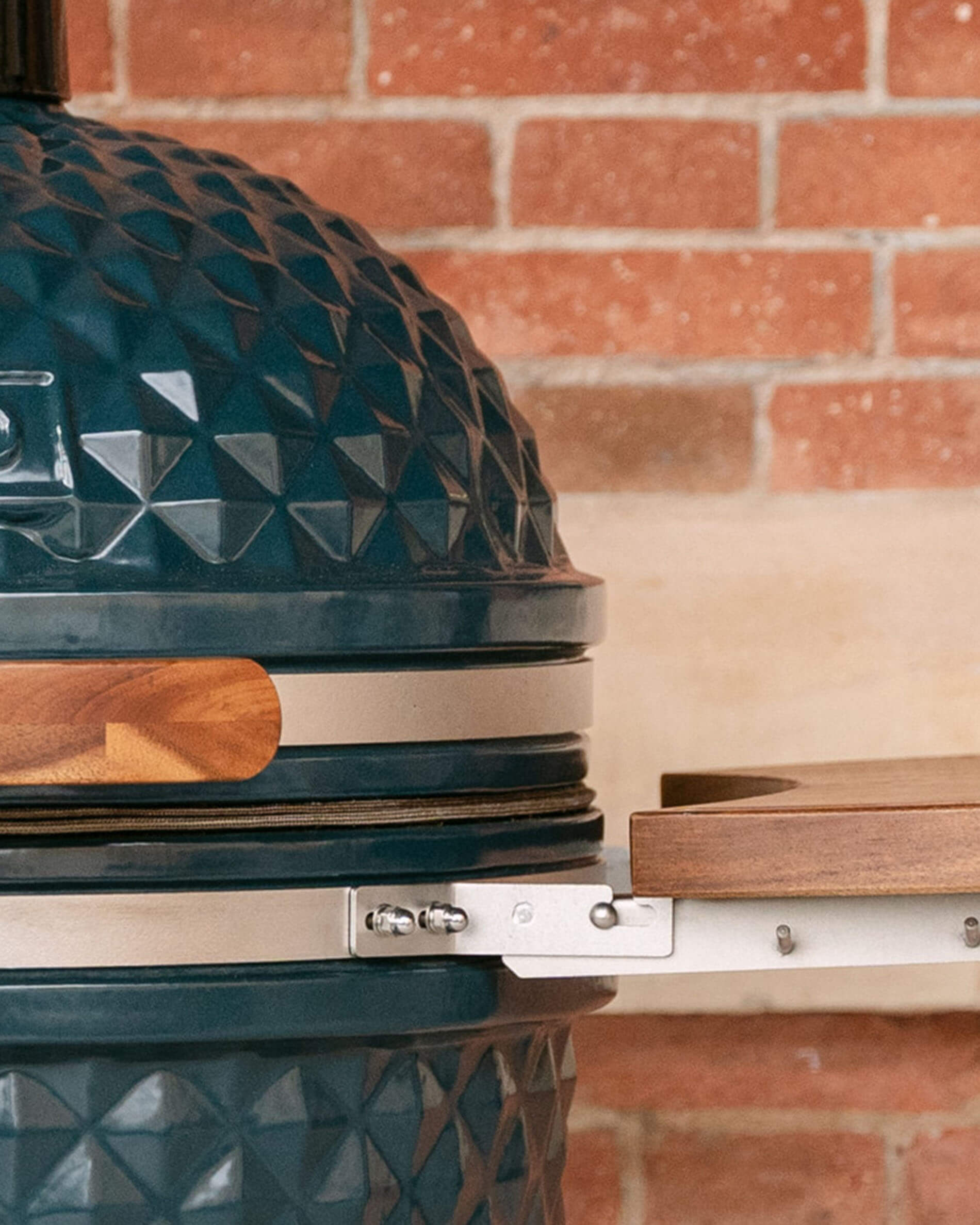 Detail der Metallbefestigungen, die eine seitliche Holzablage mit dem Kamado-Korpus verbinden, mit sichtbaren Schrauben und sichtbarer Dichtung.