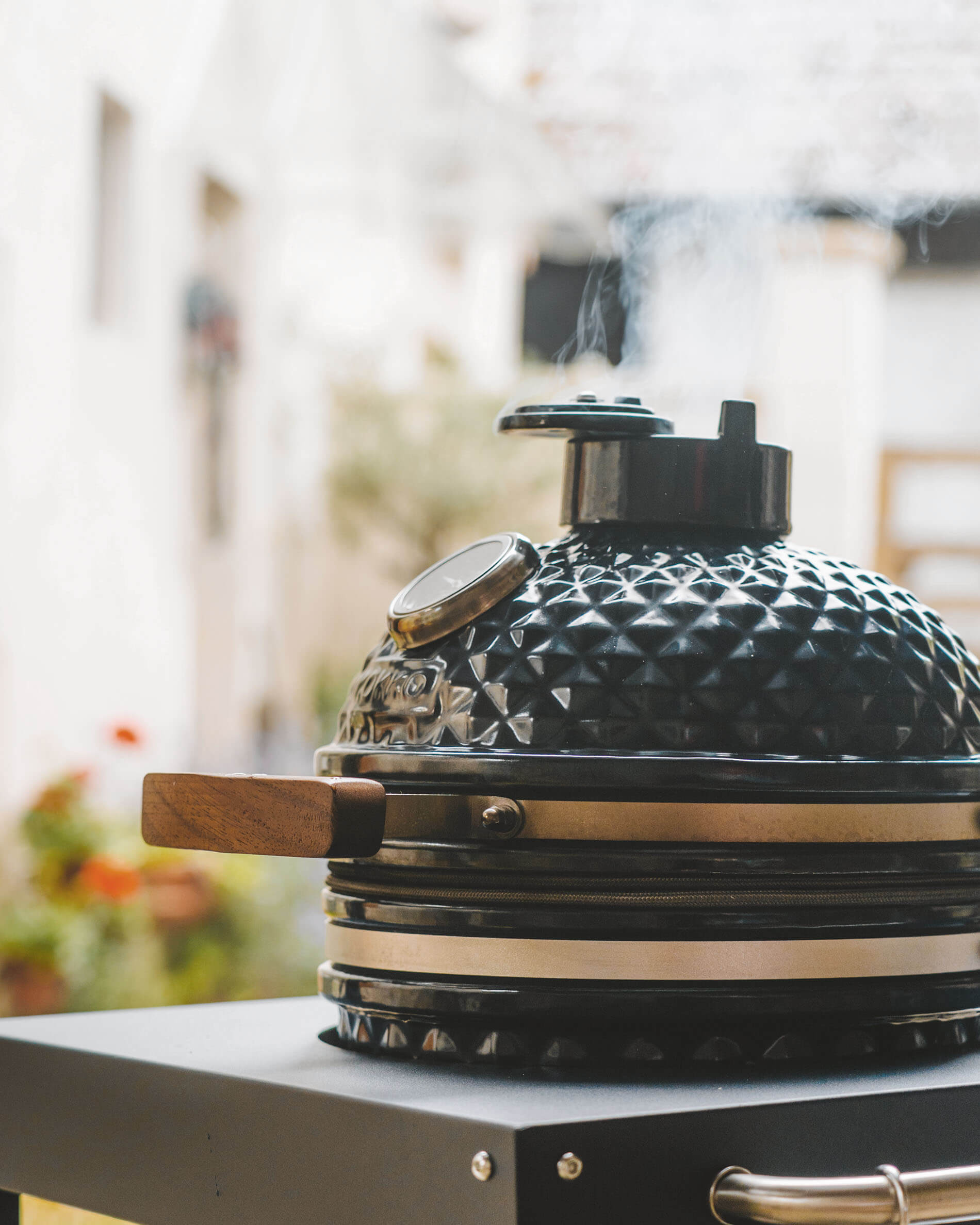 Schwarzer Kamado Kokko auf einem Wagen, mit texturiertem Keramikdeckel, Holzgriff, integriertem Thermometer und Rauch, der aus dem Kamin aufsteigt.