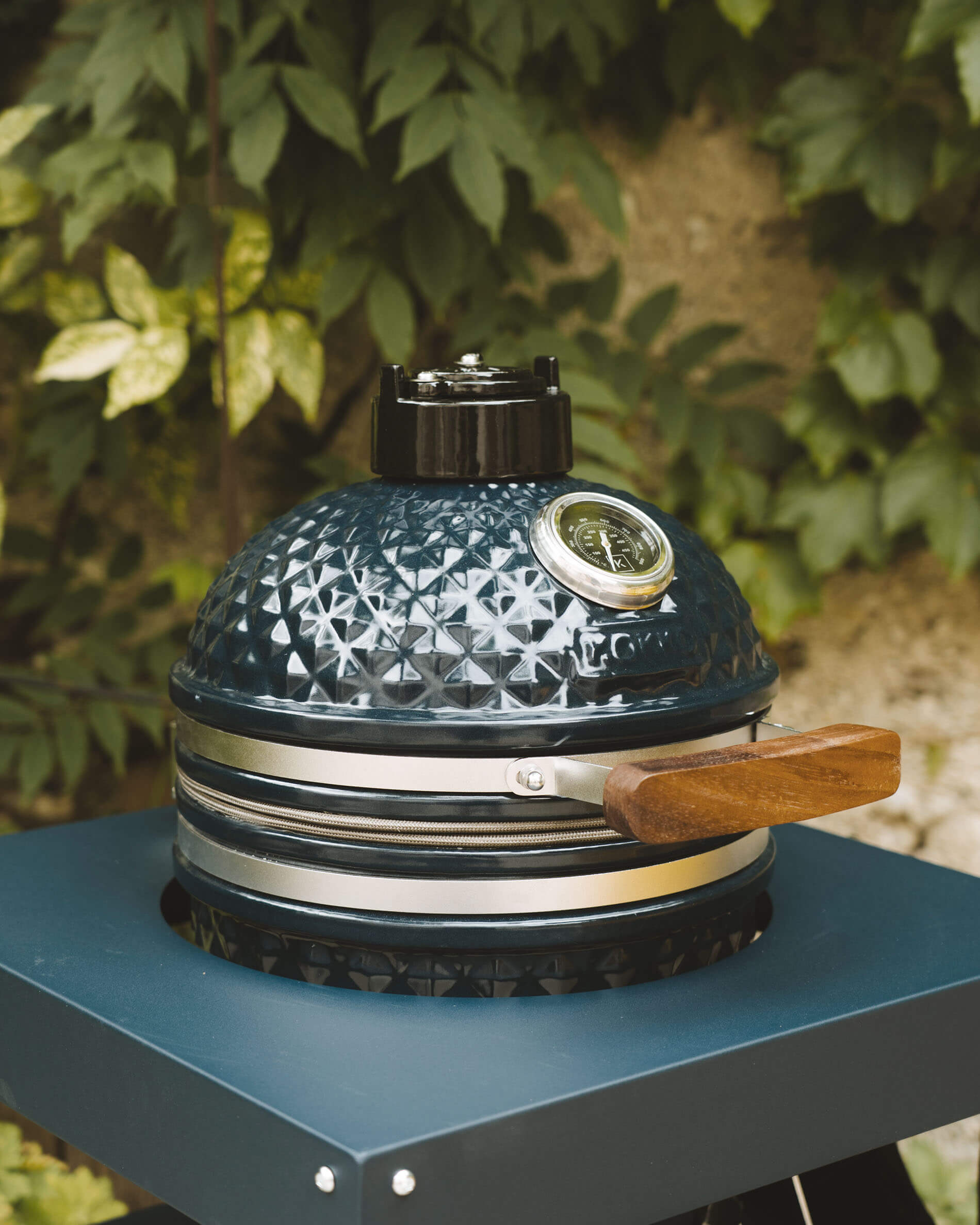 Frontansicht eines Kokko Kamado in Ozeanblau auf einem Wagen, mit Fokus auf die strukturierte Keramik, das integrierte Thermometer und den Holzgriff.