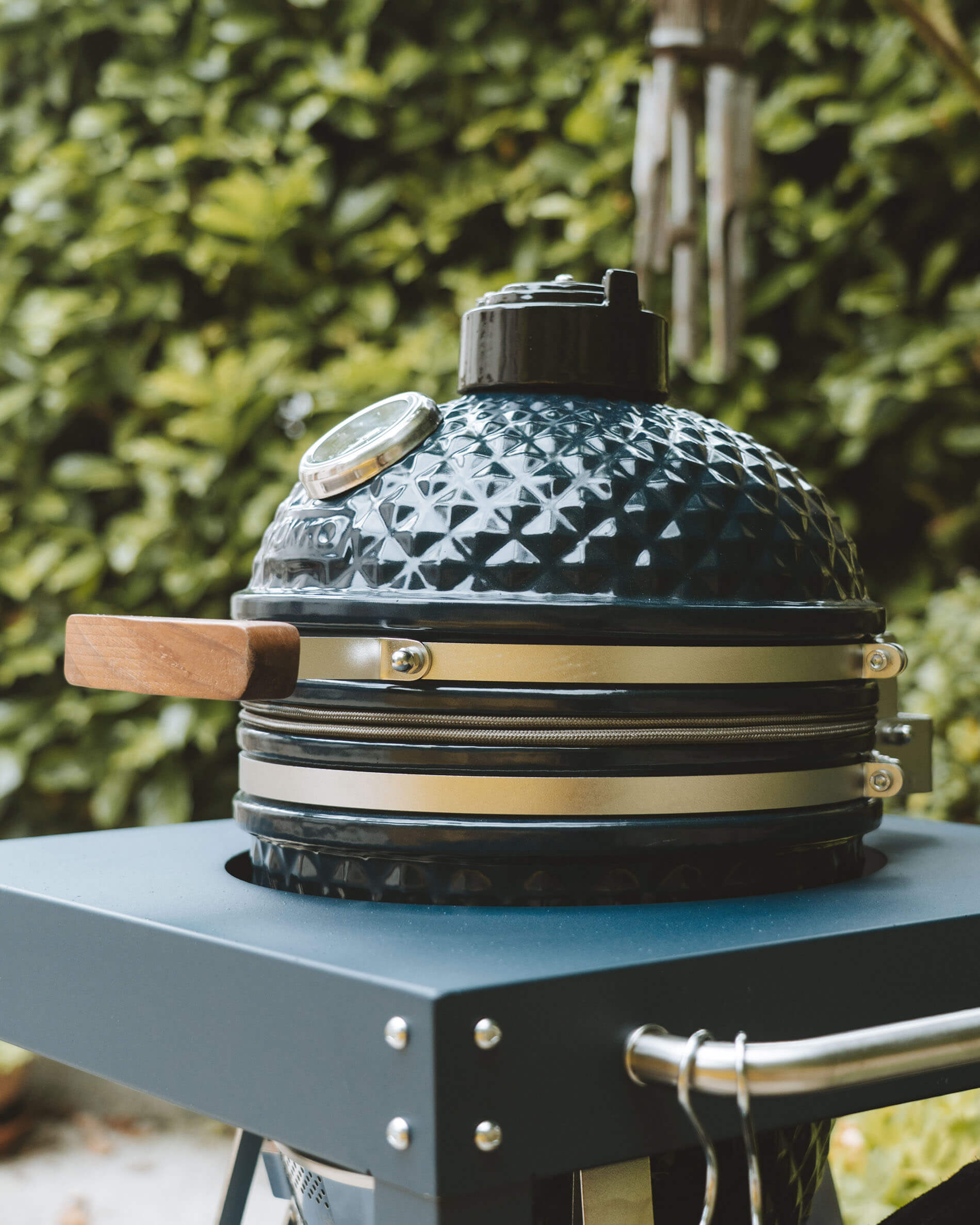 Seitenansicht eines Kokko Kamado in Ozeanblau auf einem Wagen, mit Fokus auf den Keramikdeckel, den Holzgriff und das integrierte Thermometer.