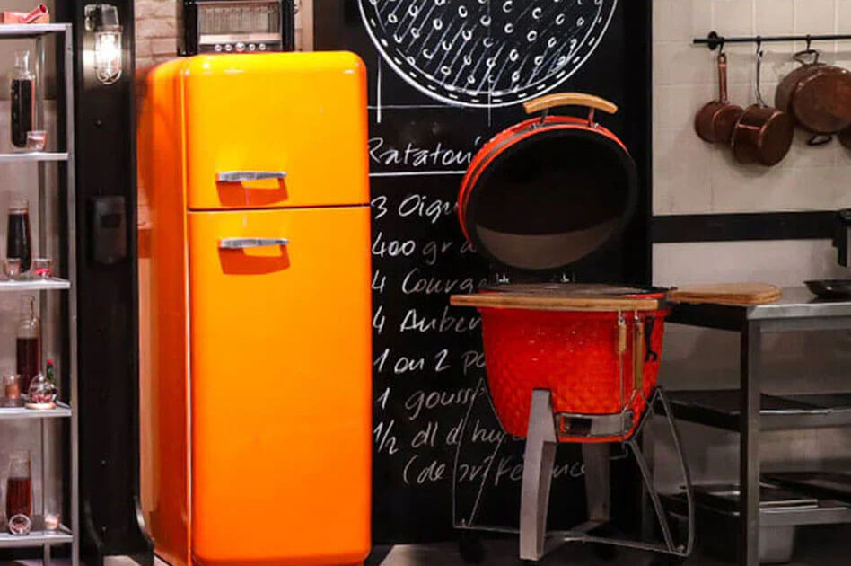 Roter Kamado Kokko aus Keramik, auf dem Set der Sendung Top Chef installiert, mit geöffnetem Deckel und seitlichen Holzablagen, in einer professionellen Studioküche.